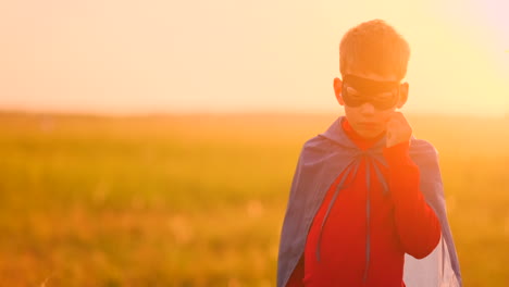 A-boy-dressed-as-a-super-hero-standing-in-a-mask-and-a-red-cloak-watching-the-sunset-in-the-field-in-the-summer.-Summer-night-the-boy-dreams-and-heroic-deeds-and-comic-books
